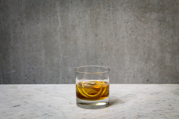 An amber coloured cocktail with an orange peel garnish in a rocks glass . The cocktail is placed on white marble and is set against a dark concrete wall.