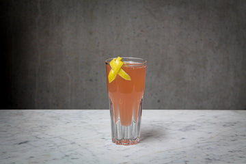 A pale pink coloured cocktail with a lemon peel garnish in an amaro glass. The cocktail is placed on white marble and is set against a dark concrete wall.