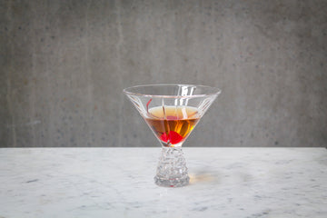 A pale amber coloured cocktail with a red cherry garnish in a martini glass . The cocktail is placed on white marble and is set against a dark concrete wall.