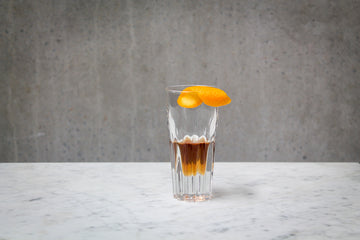 A dark orange coloured drink in an aperitif glass.  An orange twist is resting on top of the glass. The cocktail is placed on white marble and is set against a dark concrete wall.