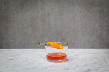 A dark orange coloured cocktail in a short glass.  A brass swizzle stick with an orange twist wrapped around it is resting across the glass. The cocktail is placed on white marble and is set against a dark concrete wall.