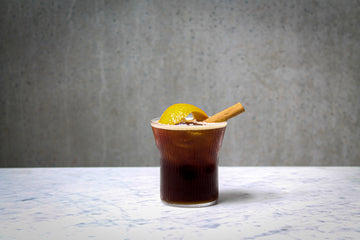 A coffee coloured cocktail in a ribbed rocks glass, garnished with a cinnamon stick and orange peel. The cocktail is placed on white marble and is set against a dark concrete wall.