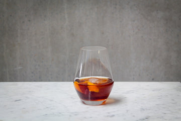 A deep amber coloured cocktail with ice and an orange peel garnish in a stemless wine glass. The cocktail is placed on white marble and is set against a dark concrete wall.