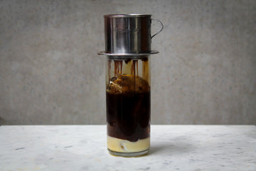 A tall glass filled with ice and a coffee coloured cocktail. There is a layer of condensed milk in the bottom, and a phin filter on top with coffee dripping through. The cocktail is placed on white marble and is set against a dark concrete wall.