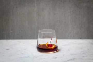 A deep amber coloured cocktail with a red cherry and orange peel garnish in a short glass. The cocktail is placed on white marble and is set against a dark concrete wall.