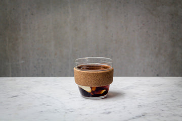 A dark brown coloured cocktail with a swirl of cream in a short coffee glass. There is a cork band around the glass. The cocktail is placed on white marble and is set against a dark concrete wall.