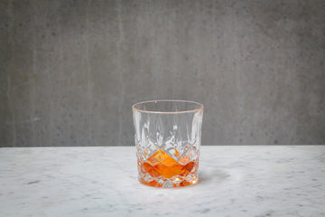 A pale orange coloured cocktail in a cut crystal rocks glass. The cocktail is placed on white marble and is set against a dark concrete wall.