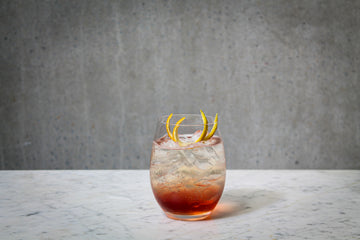 A layered clear and ruby coloured cocktail in a stemless glass, garnished with orange peel. The cocktail is placed on white marble and is set against a dark concrete wall.