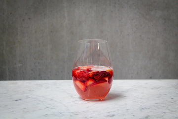 A pale pink coloured cocktail in a stemless glass, filled with cut strawberries and ice. The cocktail is placed on white marble and is set against a dark concrete wall.