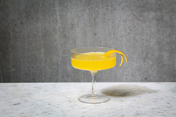 An orange coloured cocktail with an orange peel garnish in a cocktail glass . The cocktail is placed on white marble and is set against a dark concrete wall.