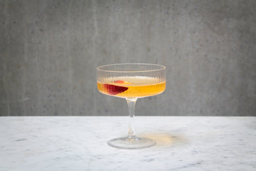 A pale orange coloured cocktail with a fresh peach garnish in a clear ribbed coupe glass. The cocktail is placed on white marble and is set against a dark concrete wall.