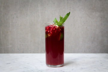 A deep red coloured cocktail with a garnish of pomegranate arils, lime wedges and a sprig of fresh mint in a tall glass. The cocktail is placed on white marble and is set against a dark concrete wall.