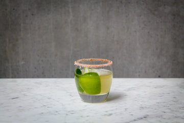 A pale green coloured cocktail with a lime wedge garnish in a short glass. There is pink salt on the rim of the glass. The cocktail is placed on white marble and is set against a dark concrete wall.
