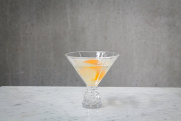 A pale lemon coloured cocktail with an orange peel garnish in a martini glass . The cocktail is placed on white marble and is set against a dark concrete wall.