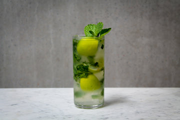 A pale green coloured cocktail with a garnish of lime wedges and fresh mint leaves in a tall glass. The cocktail is placed on white marble and is set against a dark concrete wall.