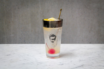 A pale lemon coloured cocktail with a lemon wedge and red cherry garnish in a highball glass with a silver band and number 5. The cocktail is placed on white marble and is set against a dark concrete wall.