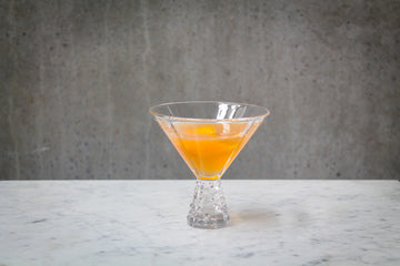 An orange coloured cocktail with an orange peel garnish in a martini glass . The cocktail is placed on white marble and is set against a dark concrete wall.