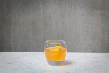 An orange coloured cocktail with a lemon peel garnish in a short glass. The cocktail is placed on white marble and is set against a dark concrete wall.
